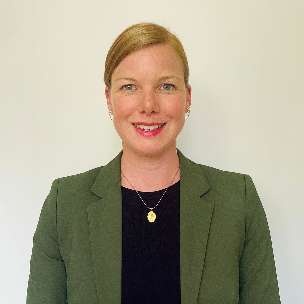 Frau mit blonden Haaren in einem grünen Blazer und schwarzem Oberteil, lächelnd vor neutralem Hintergrund.