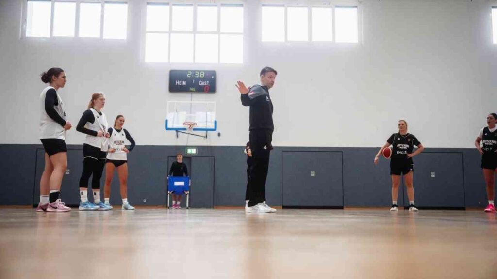 Basketballtraining mit einer Gruppe von Spielerinnen und einem Trainer in einer Sporthalle.