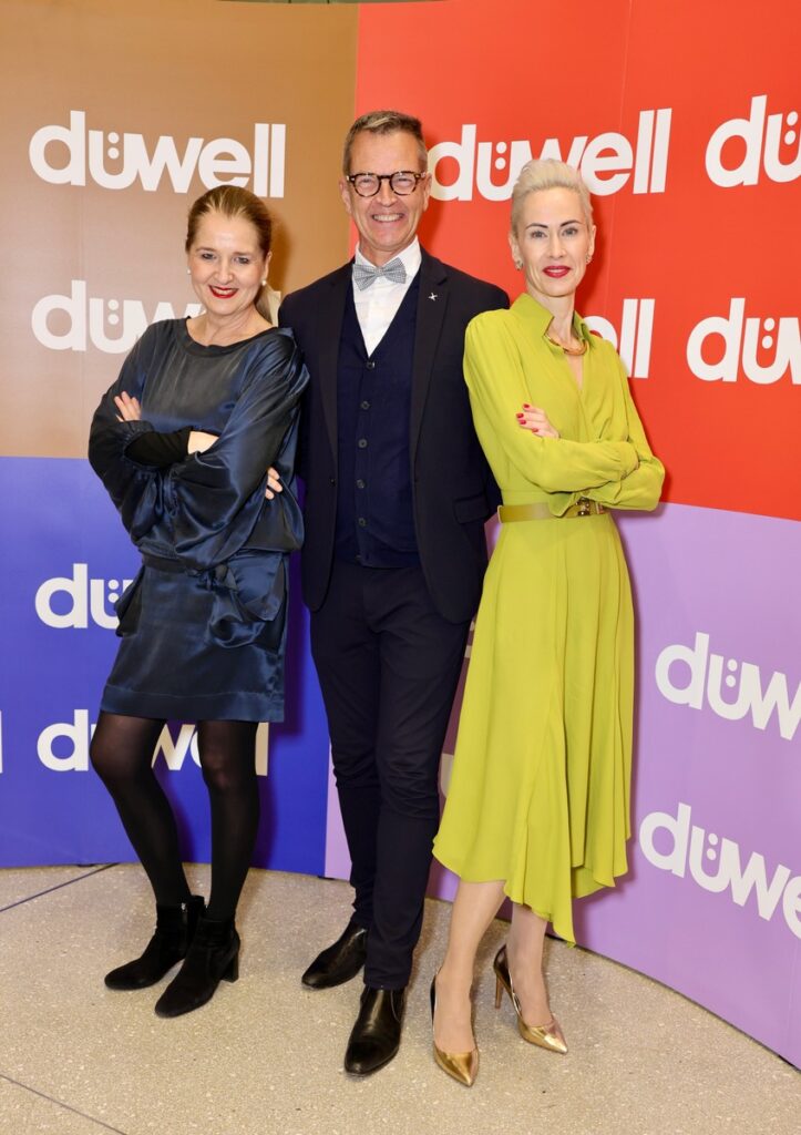 Drei Personen posieren vor einer bunten Wand mit dem Schriftzug 'duwell' in verschiedenen Farben.