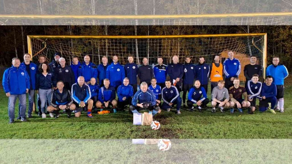 Gruppe von Fußballtrainern und Spielern in blauen Trikots vor einem Fußballtor auf dem Platz.