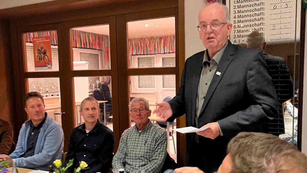 Ein Mann spricht während einer Versammlung, umgeben von mehreren Zuhörern in einem Innenraum.