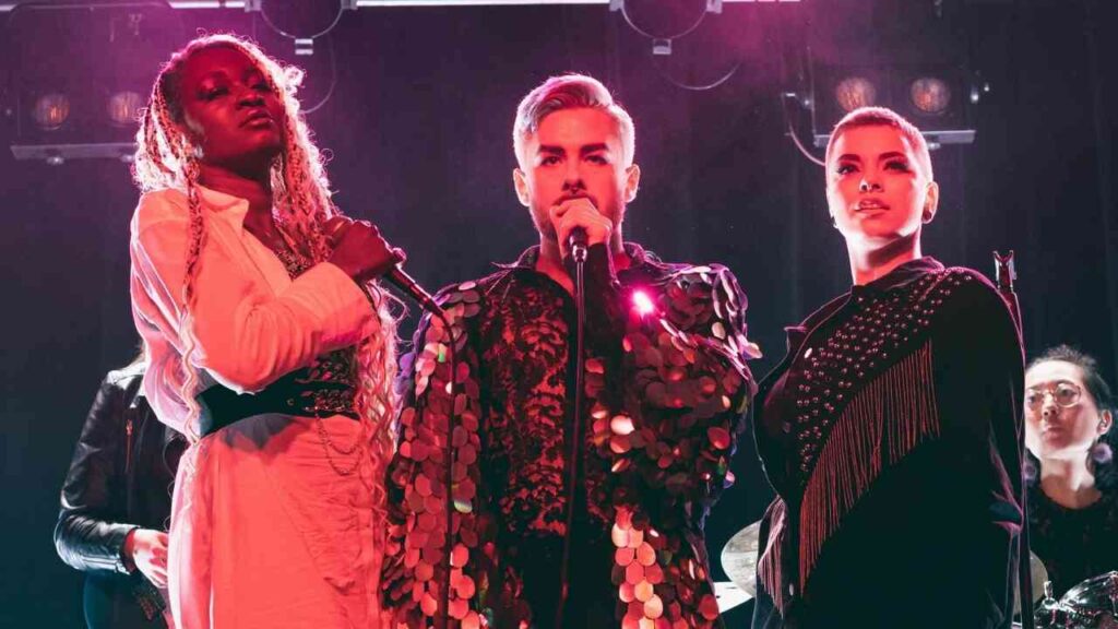 Drei Sänger auf der Bühne während eines Live-Auftritts mit Mikrofonen in der Hand und bunten Lichtern im Hintergrund.