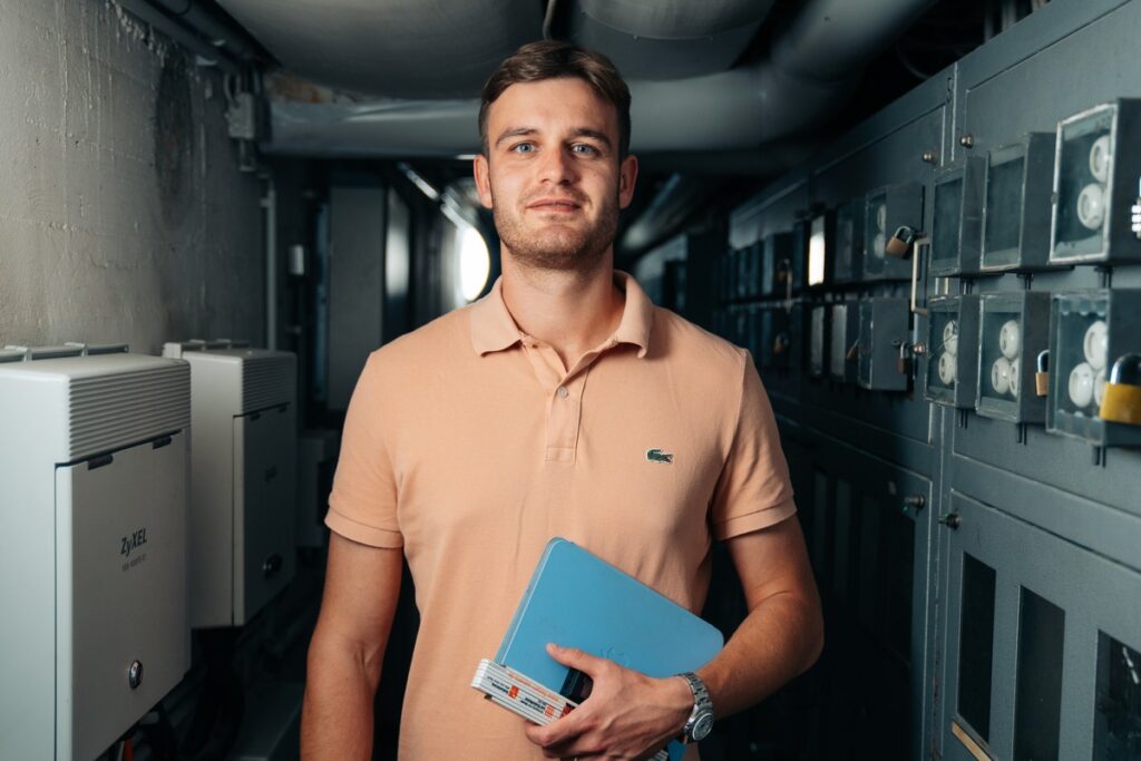 Mann in einem engen Raum mit elektrischen Schaltkästen, hält einen blauen Ordner und steht in einem professionellen Umfeld.
