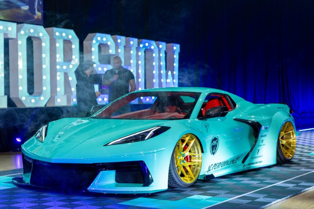 Türkisfarbener Sportwagen mit goldenen Felgen auf einer Messebühne mit beleuchtetem Schriftzug 'Motorshow'.