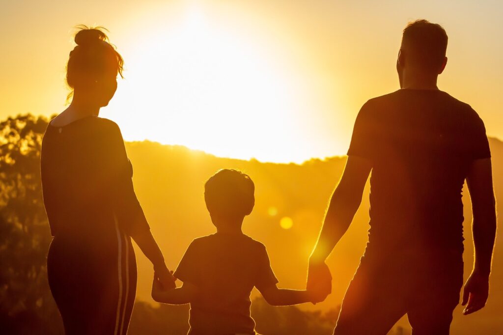 Familie, bestehend aus einem Erwachsenen, einem Kind und einer weiteren Person, die bei Sonnenuntergang Hand in Hand stehen.