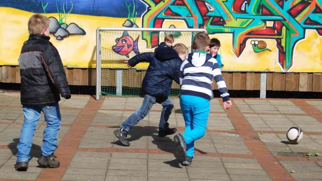 Fünf Kinder spielen Fußball auf einem Platz mit Graffiti im Hintergrund.