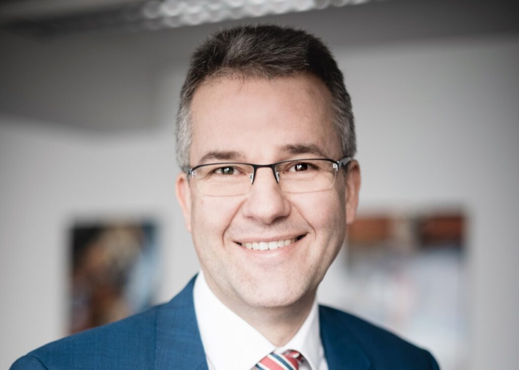 Lächelnder Mann mit Brille und Anzug, der in die Kamera schaut, mit einem freundlichen Ausdruck im Gesicht.