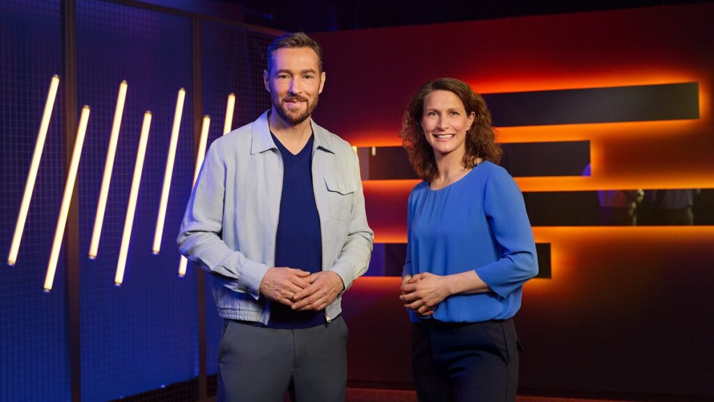 Zwei Moderatoren stehen in einem Studio mit bunten Lichtern und geometrischen Formen im Hintergrund.