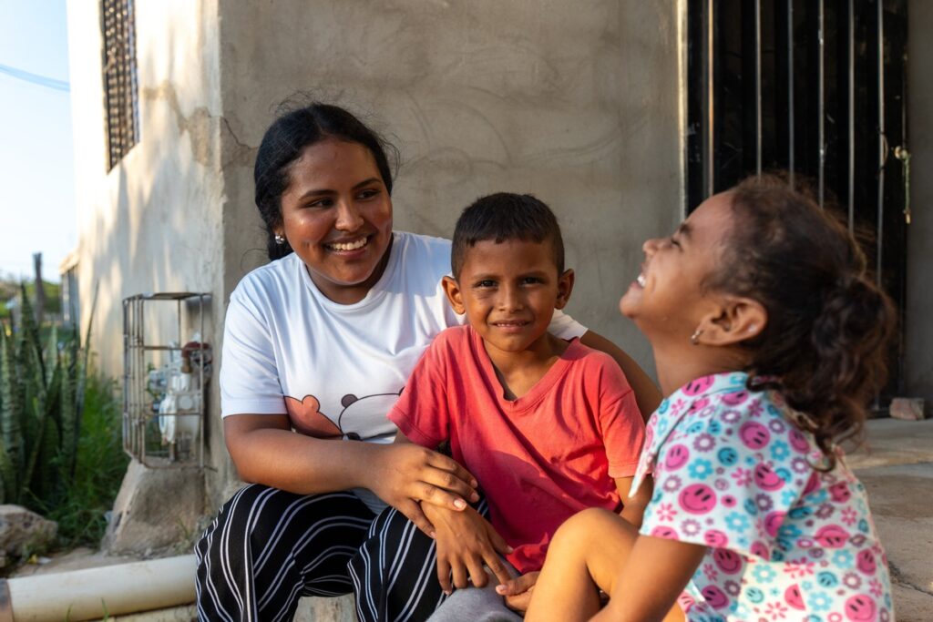 Frau sitzt mit zwei Kindern im Freien, lächelnd und interagierend, vor einem Gebäude mit einer einfachen Struktur.