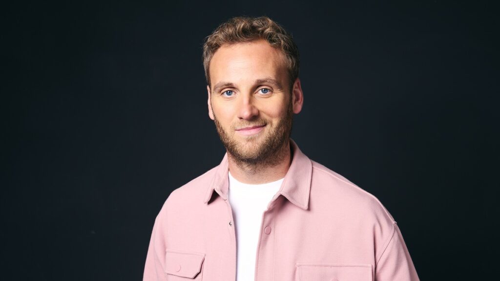 Mann mit blonden Haaren und blauem Hemd, der in die Kamera lächelt, vor einem dunklen Hintergrund.