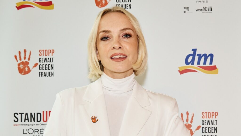 Frau mit blonden Haaren in einem weißen Anzug, die vor einem Banner mit dem Slogan 'Stopp Gewalt gegen Frauen' steht.