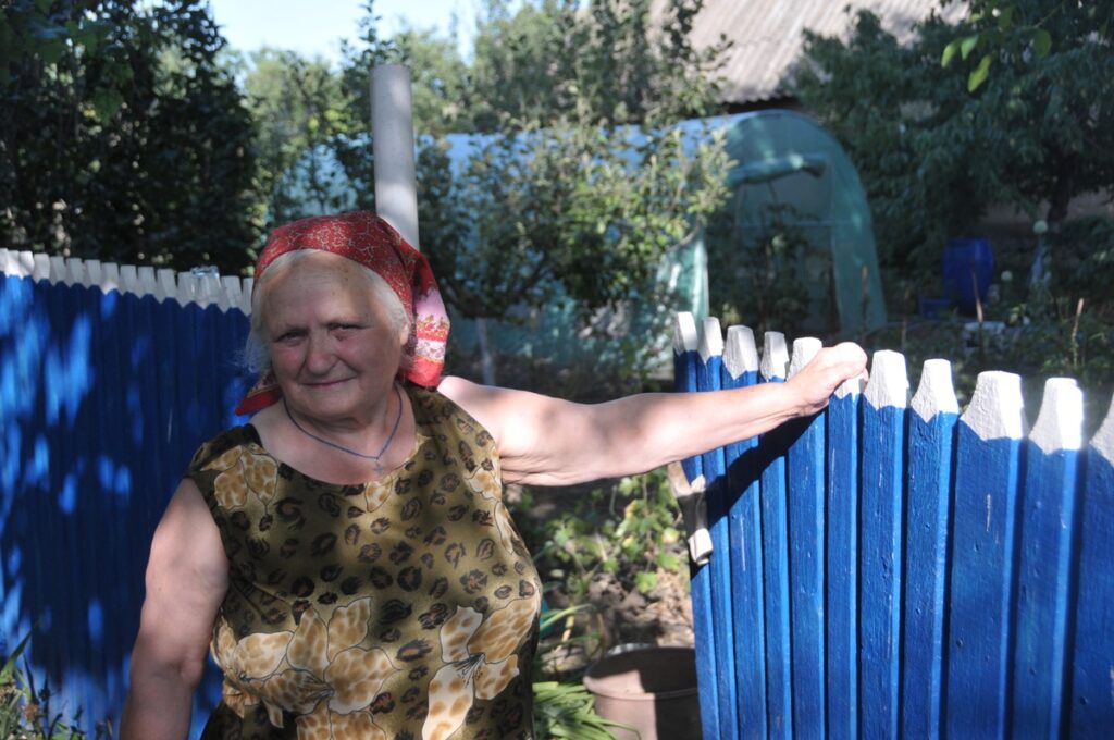 Ältere Frau mit Kopftuch steht an einem blauen Holzzaun in einem Garten.