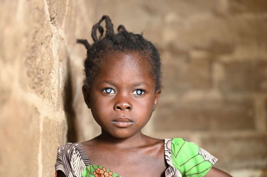 Ein Mädchen mit dunkler Haut und Zöpfen, das in einem bunten Kleid mit grünem Muster in die Kamera schaut.