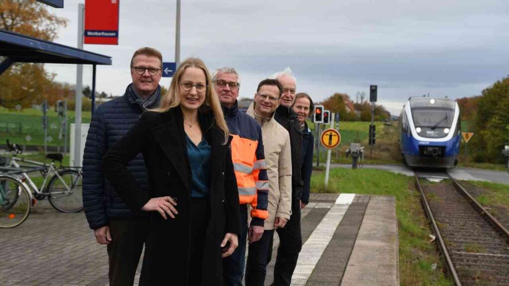 Gruppe von sechs Personen, die an einem Bahnhof stehen, während ein Zug vorbeifährt.