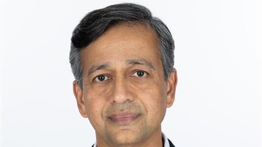 Porträt eines Mannes mit grauen Haaren und einem neutralen Gesichtsausdruck vor einem weißen Hintergrund.