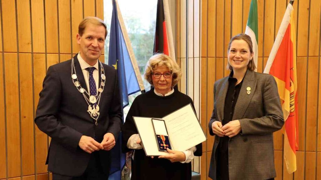 Drei Personen stehen in einem Raum, eine Frau hält eine Auszeichnung in der Hand, während zwei weitere Personen daneben stehen.