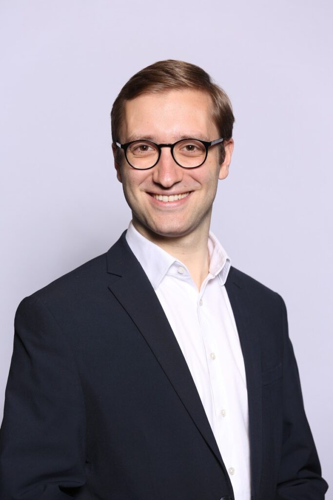 Mann mit Brille und Anzug, der freundlich in die Kamera lächelt, vor einem neutralen Hintergrund.