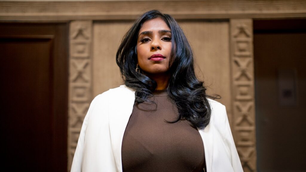 Frau mit langen, schwarzen Haaren und braunem Oberteil, die in einem eleganten weißen Blazer posiert.