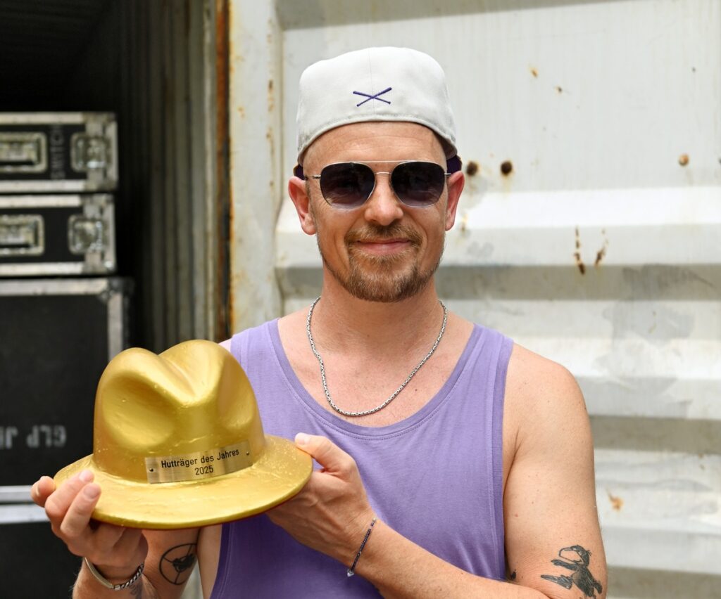 Mann mit einer goldenen Auszeichnung in Form eines Huts, trägt ein lila Oberteil und eine weiße Mütze.