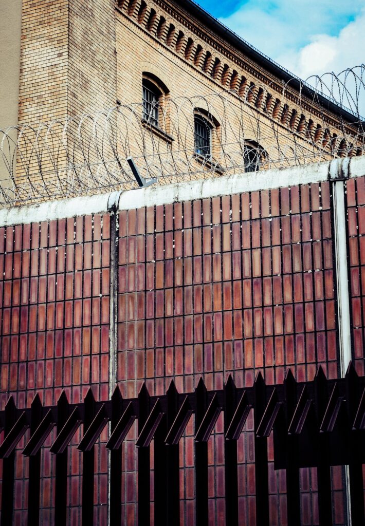 Mauer mit Stacheldraht und Ziegelsteinen, die ein Gefängnisgebäude im Hintergrund umgibt.
