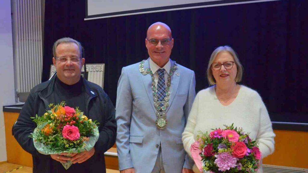 Drei Personen stehen auf einer Bühne, zwei mit Blumensträußen und einer in einem Anzug mit einer Kette.