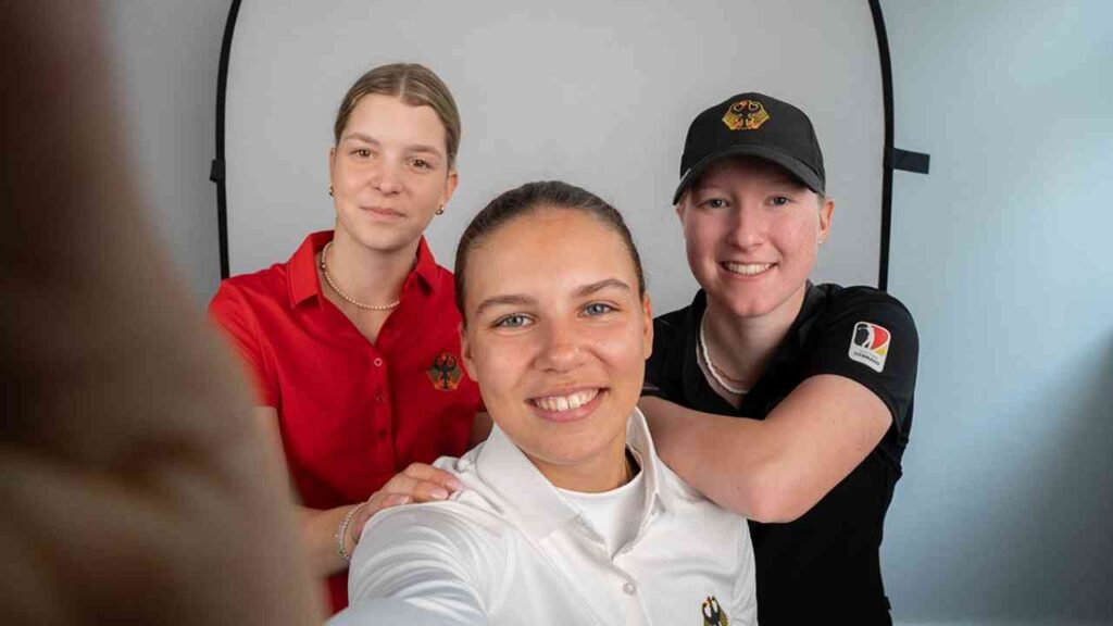Drei junge Frauen posieren für ein Selfie, eine trägt ein rotes Hemd, die andere ein weißes und die dritte ein schwarzes Hemd mit einem Logo.