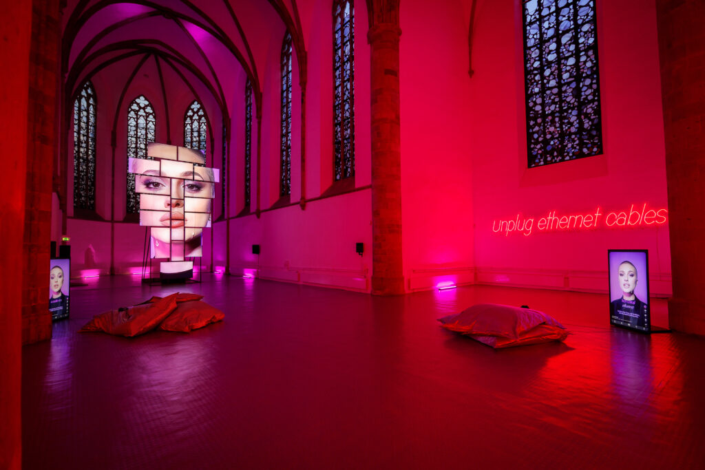 Installation in einem Raum mit pinker Beleuchtung, die ein großes Gesicht auf einer Wand zeigt und Kissen auf dem Boden platziert.