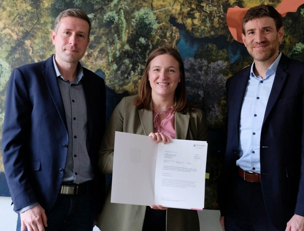 Drei Personen stehen zusammen und halten ein Dokument in der Hand, vor einer farbigen Wand mit Naturmotiv.