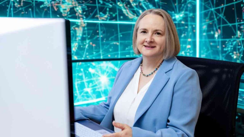Frau mit blauer Jacke sitzt vor einem Computer und lächelt, Hintergrund mit digitalen Grafiken.