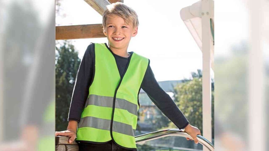 Junge mit leuchtend gelber Warnweste und reflektierenden Streifen, lächelnd und stehend an einem Geländer.