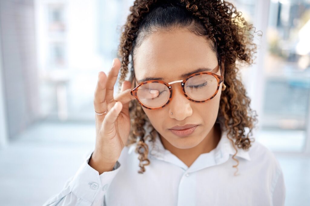 Frau mit lockigem Haar und Brille, die ihre Schläfen mit der Hand berührt und einen nachdenklichen Ausdruck zeigt.
