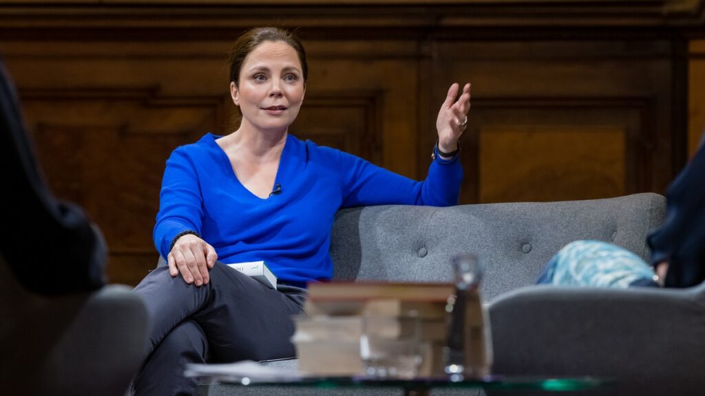 Frau in blauer Bluse sitzt auf einem grauen Sofa und spricht während einer Diskussion auf einer Bühne.