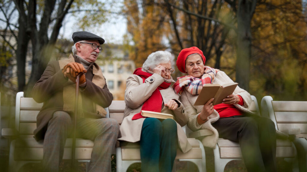 Drei ältere Menschen sitzen auf einer Bank im Park und lesen Bücher, umgeben von herbstlichen Bäumen.
