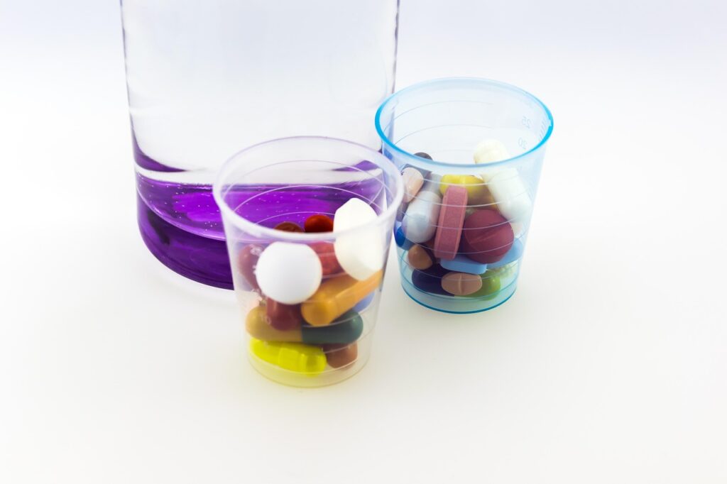 Zwei transparente Becher mit verschiedenen Tabletten und Kapseln, neben einer Flasche mit lila Flüssigkeit.