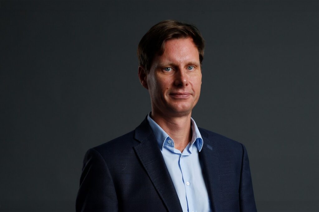 Mann in Anzug mit blauer Hemd und neutralem Hintergrund, steht aufrecht und schaut direkt in die Kamera.