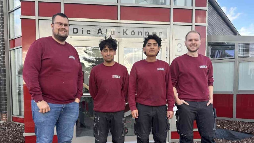 Vier Fachkräfte in roten Sweatshirts vor dem Eingang eines Unternehmens mit dem Namen 'Die Alu-Könner'.