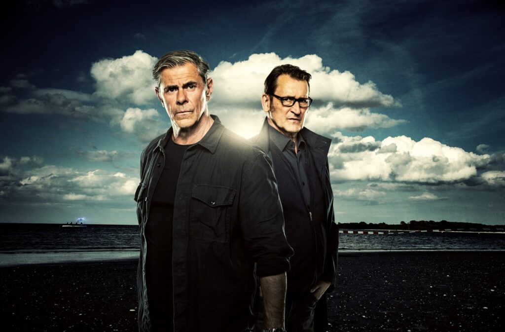 Zwei Männer in dunkler Kleidung stehen vor einer dramatischen Wolkenlandschaft am Strand.