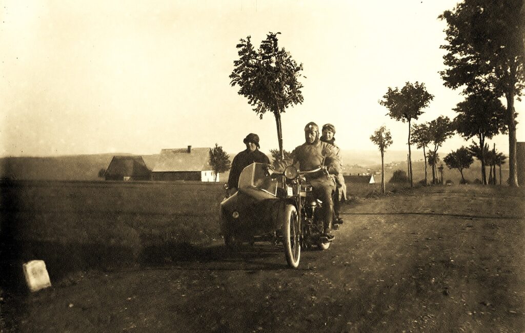 Drei Personen auf einem Motorrad mit Beiwagen, das auf einem Feldweg fährt, umgeben von Bäumen und einem Bauernhaus.