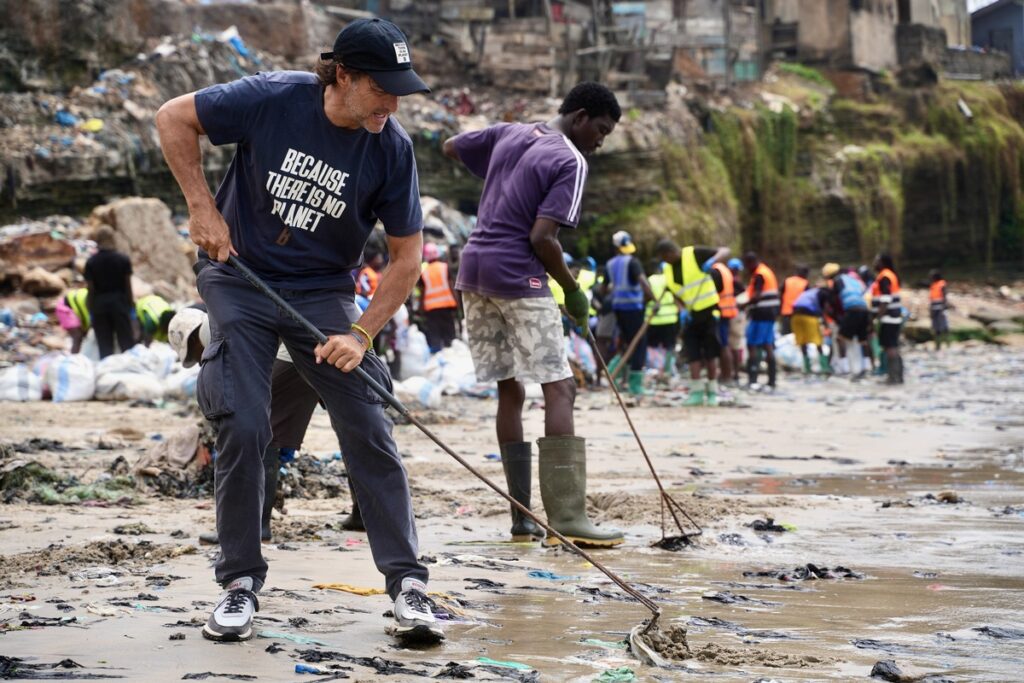 Mann mit T-Shirt, der am Strand Müll aufnimmt, während andere Helfer im Hintergrund aktiv sind.