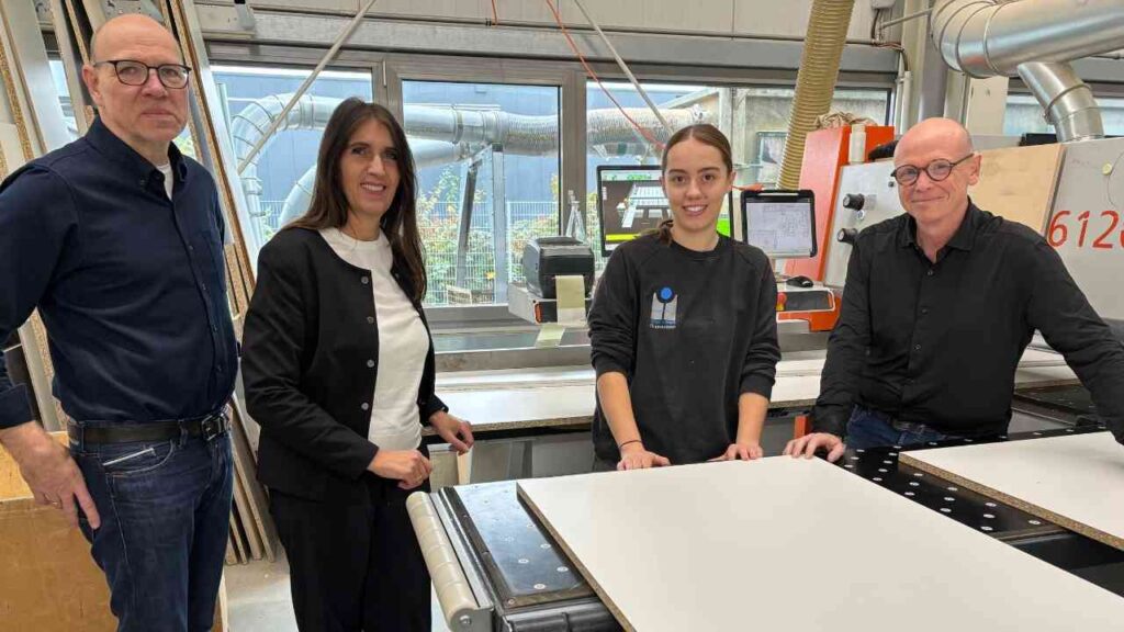 Vier Personen stehen an einer Holzverarbeitungsmaschine in einer Werkstatt, während sie Holzplatten bearbeiten.