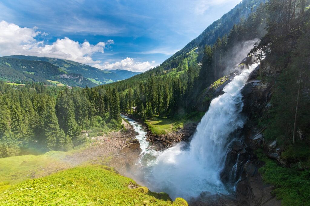 Wasserfall in einer grünen Landschaft mit Bergen und Bäumen im Hintergrund.