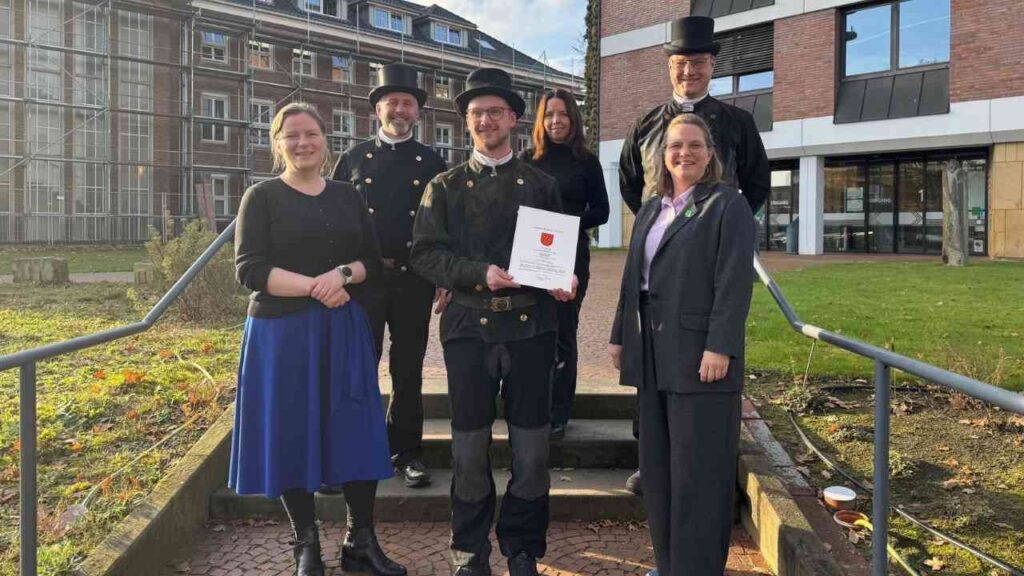 Gruppe von Personen in traditioneller Kleidung, die ein Dokument mit dem Wappen halten, vor einem modernen Gebäude.