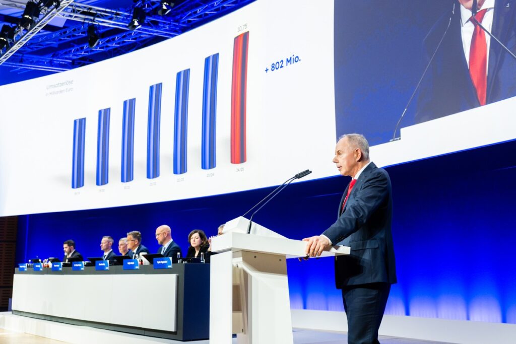 Mann am Rednerpult während einer Präsentation mit Diagramm im Hintergrund, das finanzielle Daten zeigt.