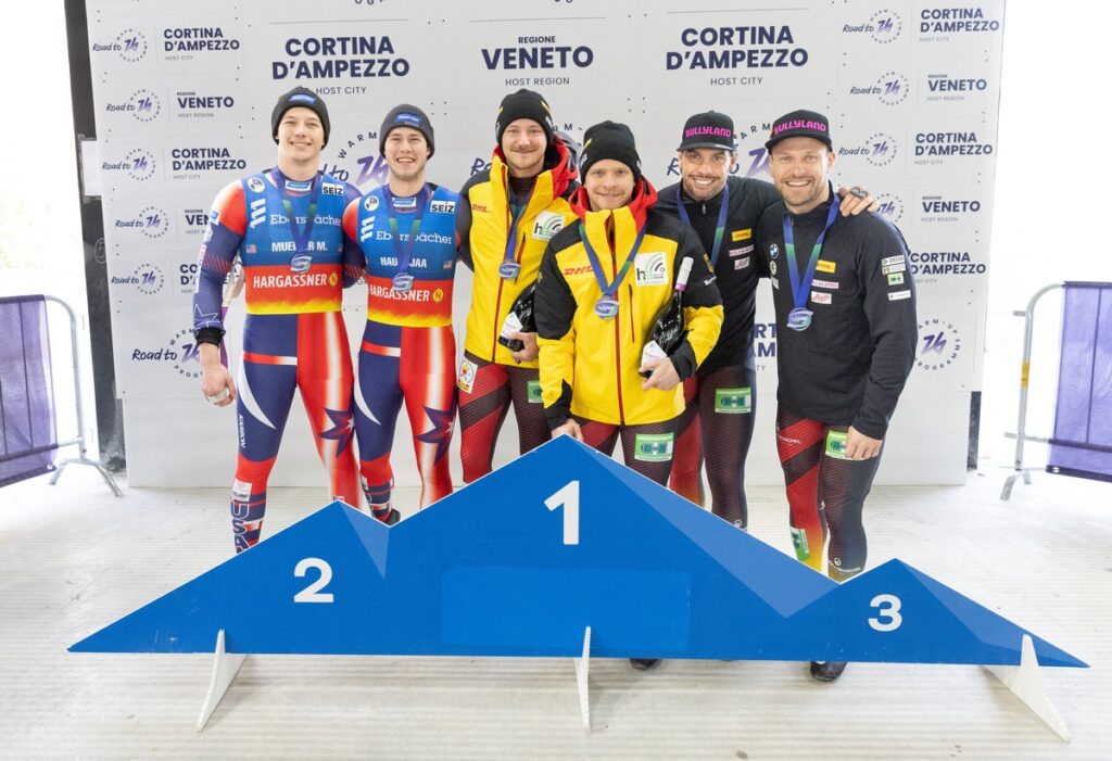 Sechs Athleten stehen auf dem Podium bei einem Wettkampf in Cortina d'Ampezzo, gekleidet in Sportanzügen mit Medaillen.