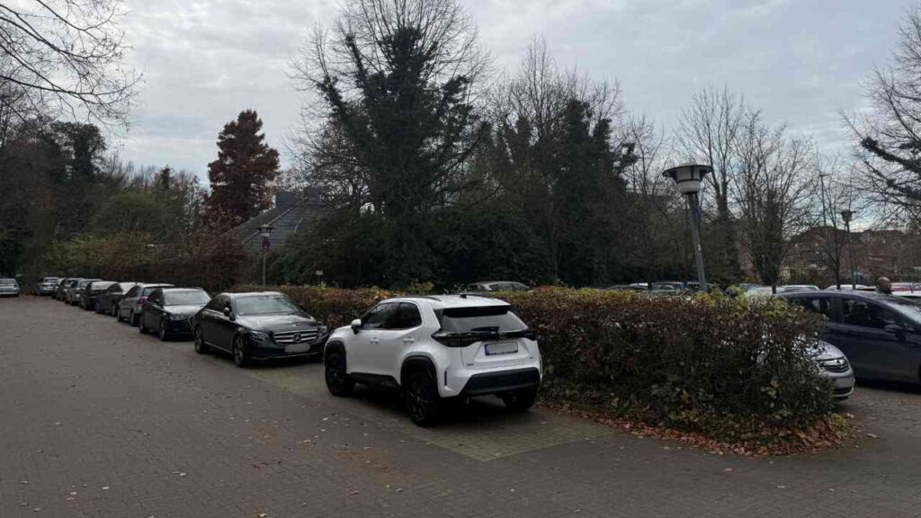 Weißes Auto parkt auf einem Parkplatz zwischen anderen Fahrzeugen in einer städtischen Umgebung.