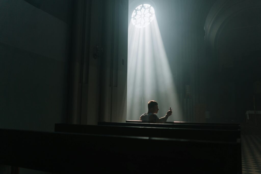 Eine Person sitzt in einer Kirche und hält eine Kerze unter einem Lichtstrahl, der durch ein Fenster fällt.