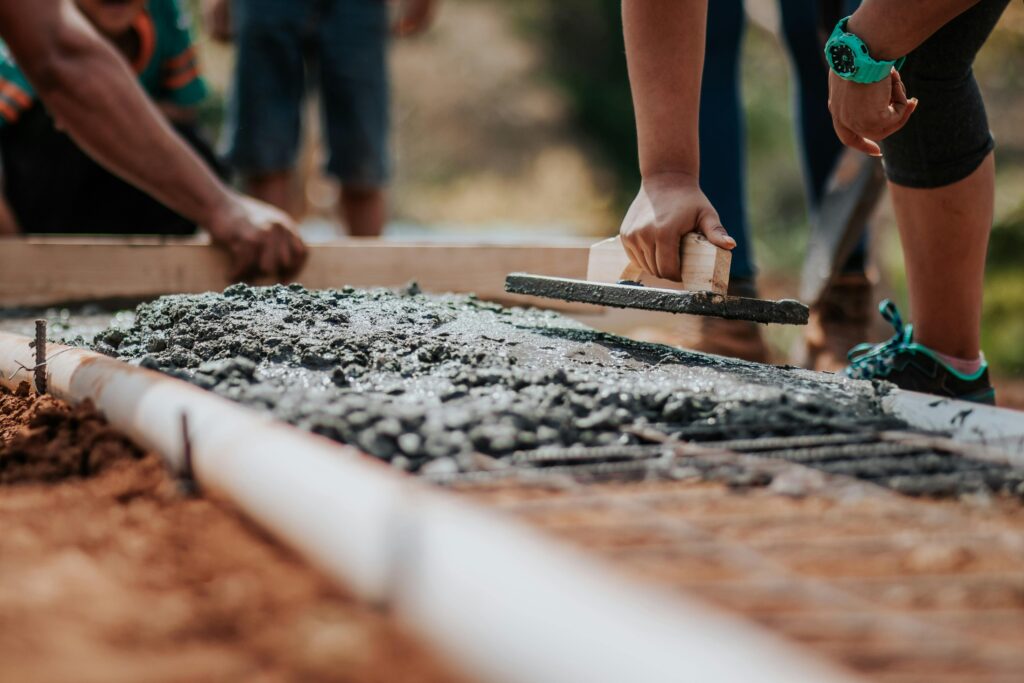 Person, die mit einem Kellenwerkzeug Beton auf einer Baustelle glättet, während andere Arbeiter im Hintergrund sichtbar sind.