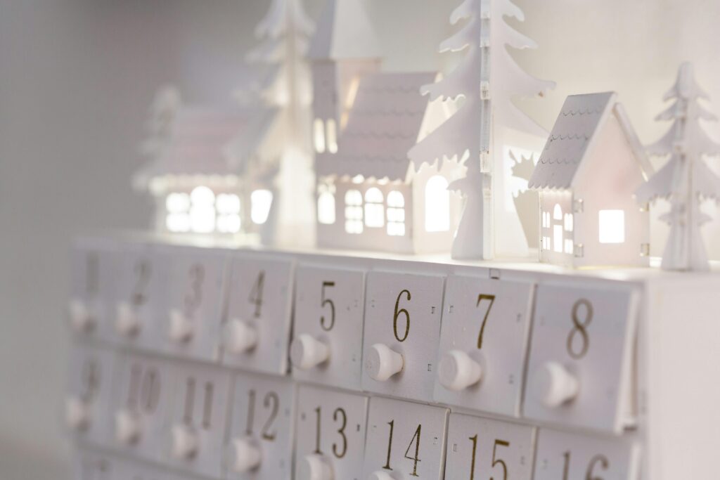 Ein Adventskalender mit weißen Häuschen und Bäumen, die sanft beleuchtet sind, auf einem weißen Hintergrund.