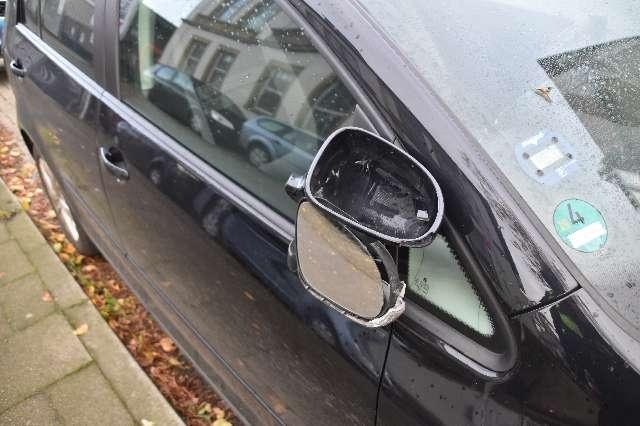 Beschädigter Seitenspiegel eines schwarzen Autos, das am Straßenrand geparkt ist.