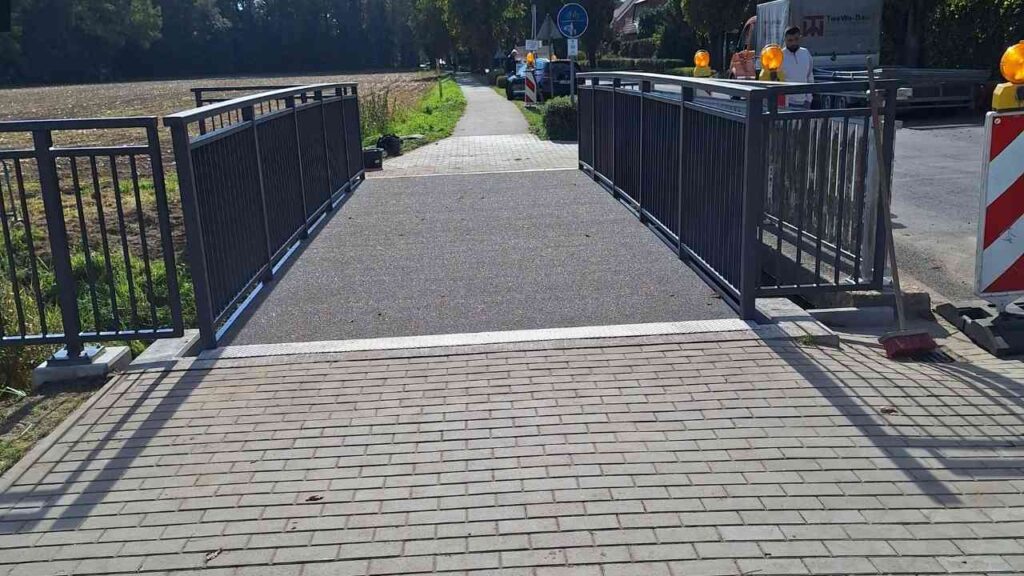 Radwegbrücke mit grauem Belag und schwarzen Geländern, die einen Weg überquert.